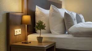Een net opgemaakt bed met witte lakens, een kleine plant in pot en een bedlamp op een houten nachtkastje.