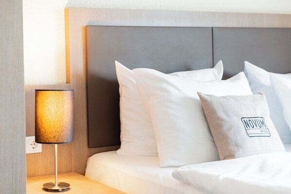 A neatly made bed with white linens and a grey pillow with "NOVUM HOTELS" logo.