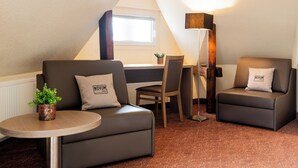 Een moderne kamer met een bank, fauteuil en een klein tafeltje met een plant. Er is een bureau met een stoel en een lamp.
