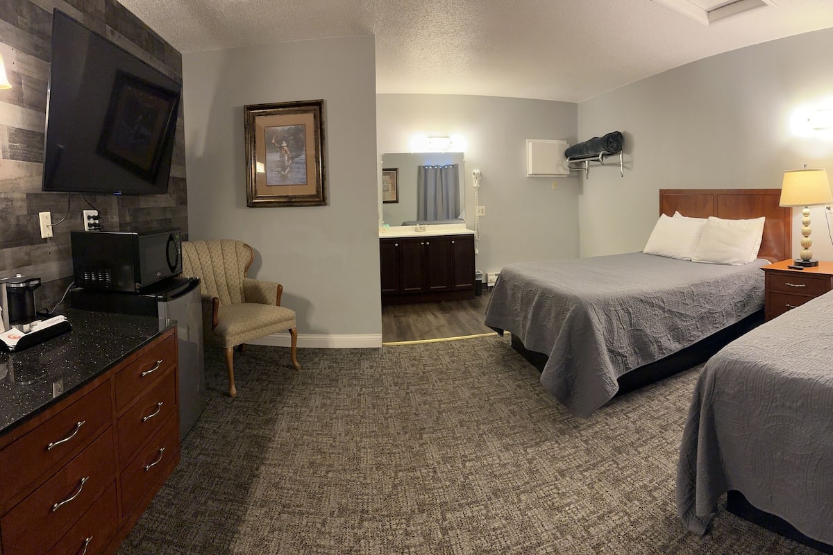 A hotel room with two beds, a TV, a desk with a chair, a dresser, and a bathroom with a mirror and sink.