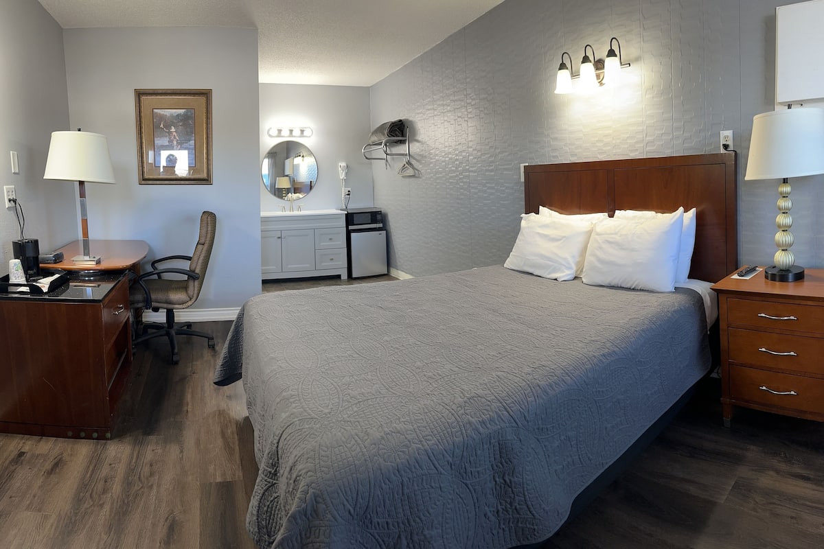 A hotel room with a large bed, a desk with a chair, a vanity with a mirror, and a nightstand with a lamp.