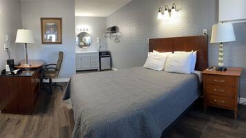 A hotel room with a large bed, a desk with a chair, a vanity with a mirror, and a nightstand with a lamp.