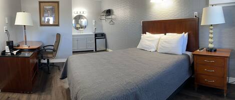 Una habitación de hotel con una cama grande, un escritorio con silla, un tocador con espejo y una mesita de noche con lámpara.