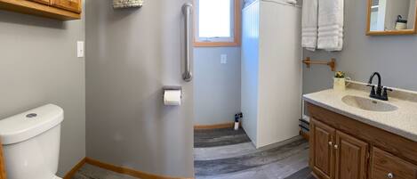 A bathroom with a toilet, a handrail, a sink with a mirror, and a window.