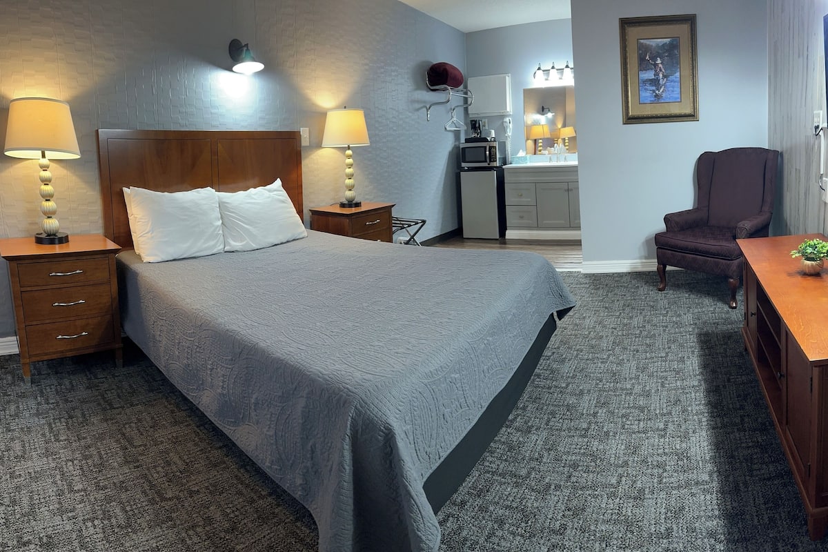 A neatly arranged hotel room with a large bed, bedside tables with lamps, a desk, a chair, a microwave, and a small kitchenette.