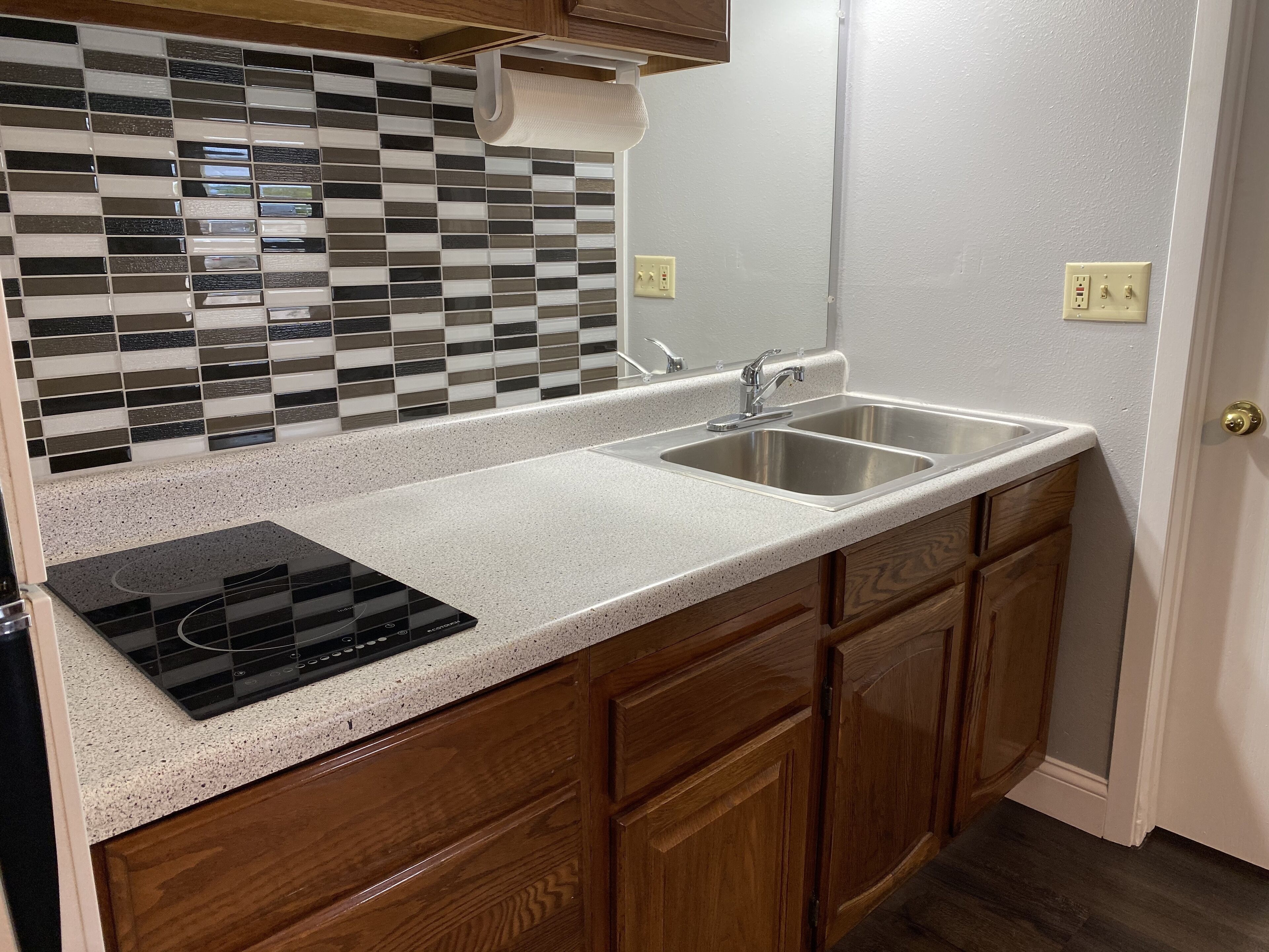 A kitchen with a black and white tiled backsplash, a stainless steel sink, a built-in stovetop, wooden cabinets, and a white countertop.