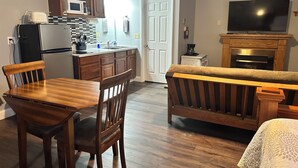 A kitchen with wooden cabinets, a refrigerator, a microwave, and a sink. There is a dining table with chairs and a sofa. A television is mounted above a fireplace. - Pelican Motel (Pelican Rapids)
