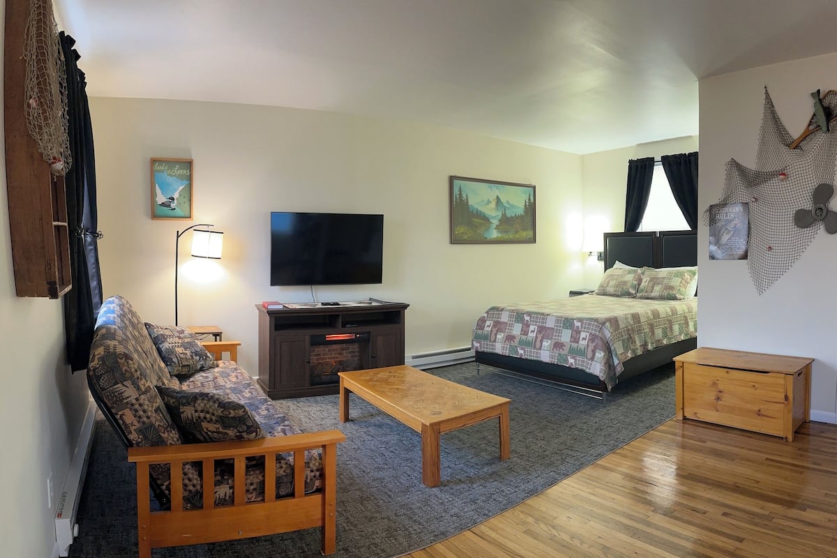 A furnished room with a sofa, a wooden table, a bed, a TV, and artwork on the walls.