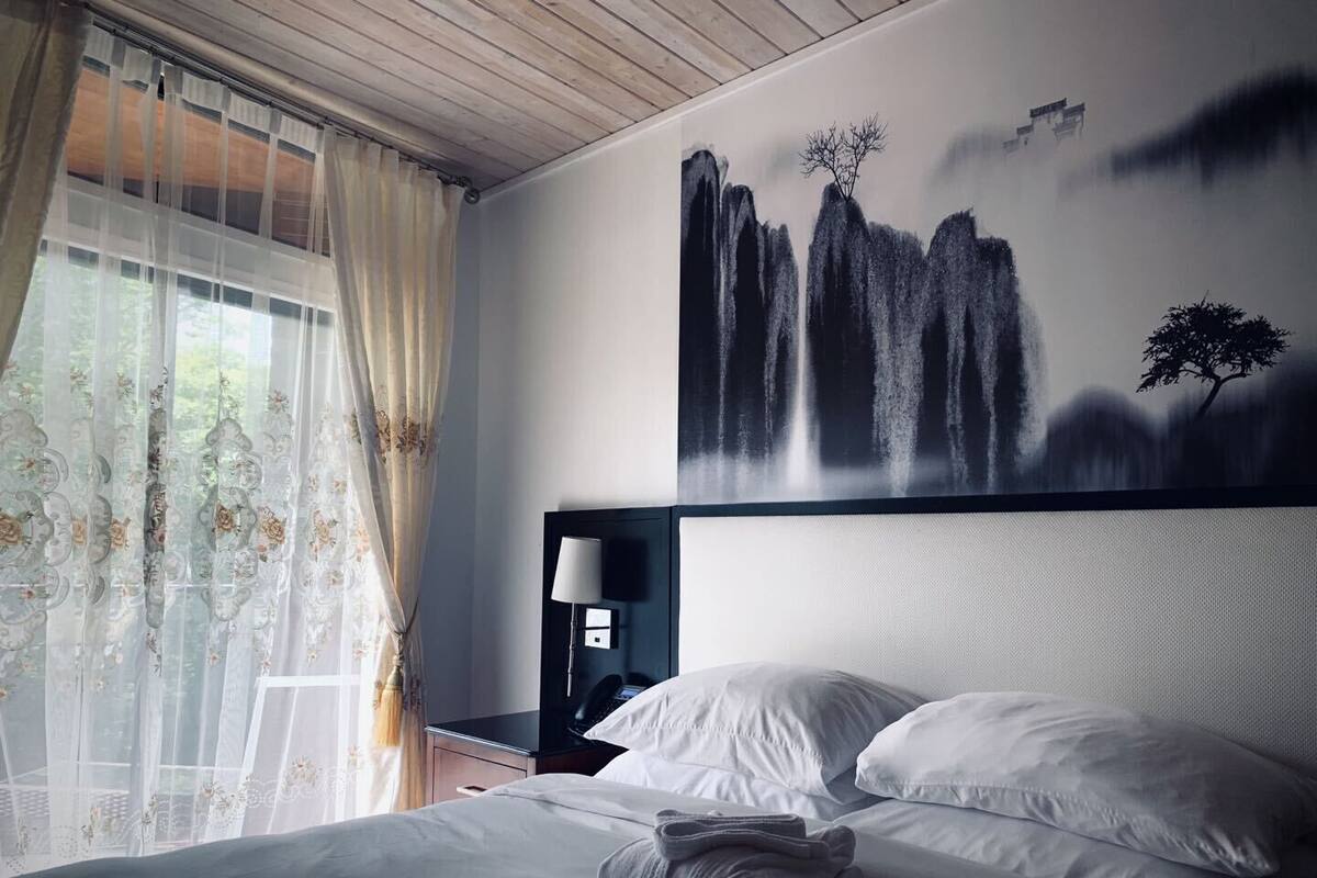 Habitación de hotel con una cama, una mesita de noche, una lámpara y un cuadro de montañas.