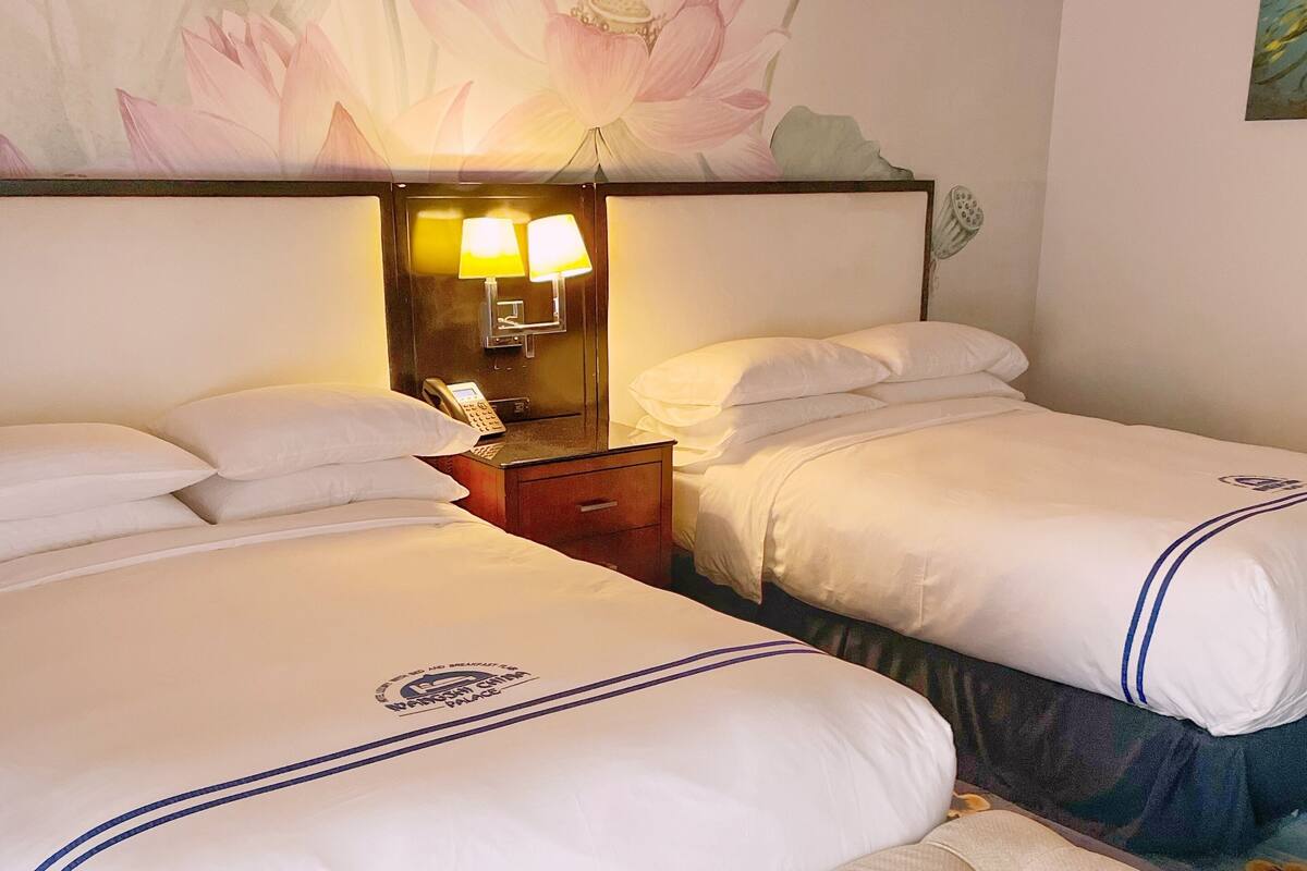 Habitación de hotel con dos camas, una mesita de noche con lámpara y un gran mural floral en la pared.