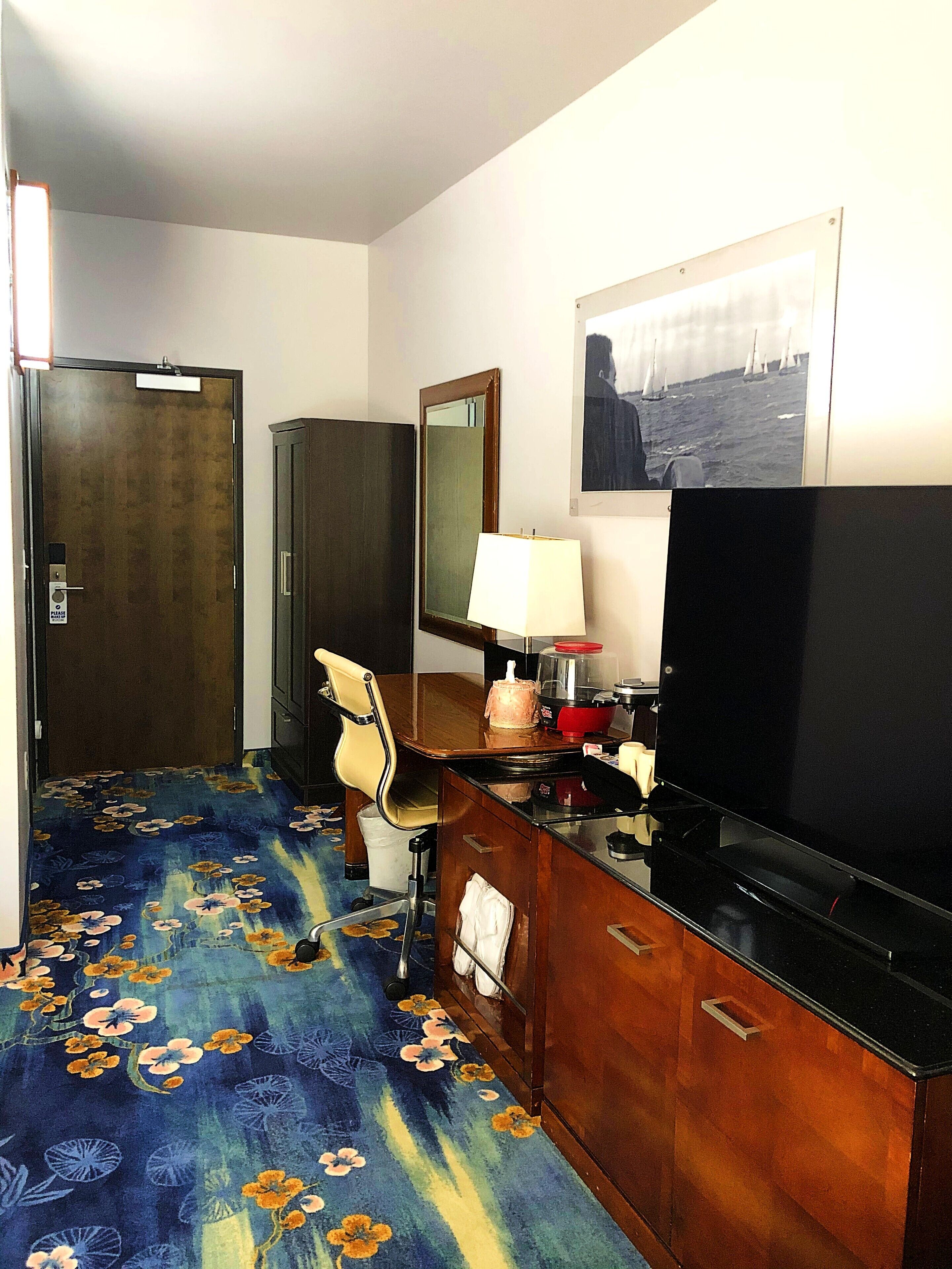 Une chambre d’hôtel avec un bureau en bois, une télévision, un miroir, une chaise et une moquette à motifs floraux.