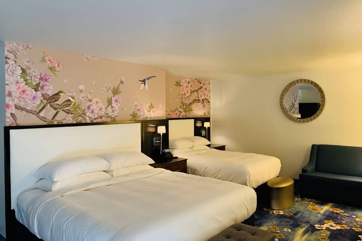 Habitación de hotel con dos camas, un sofá, un espejo redondo y decoración floral en la pared.