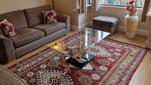 Una sala de estar con sofá, una mesa de centro, alfombra estampada, lámpara de pie y ventana con cortinas.