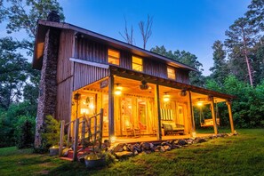 A wooden cabin with a porch, stone walls, and a well-lit interior. - Romantic StackRock Cabin – An Exclusive, Centrally Located Retreat. (Ellijay)