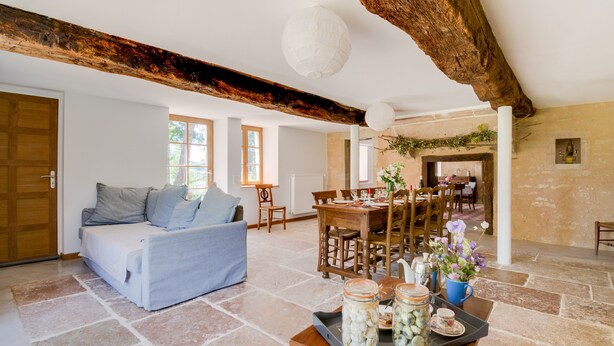 Un salón amplio con sofá, mesa de comedor y sillas. Hay frascos con alimentos en conserva sobre la mesa, y una planta en maceta en el alféizar de la ventana.