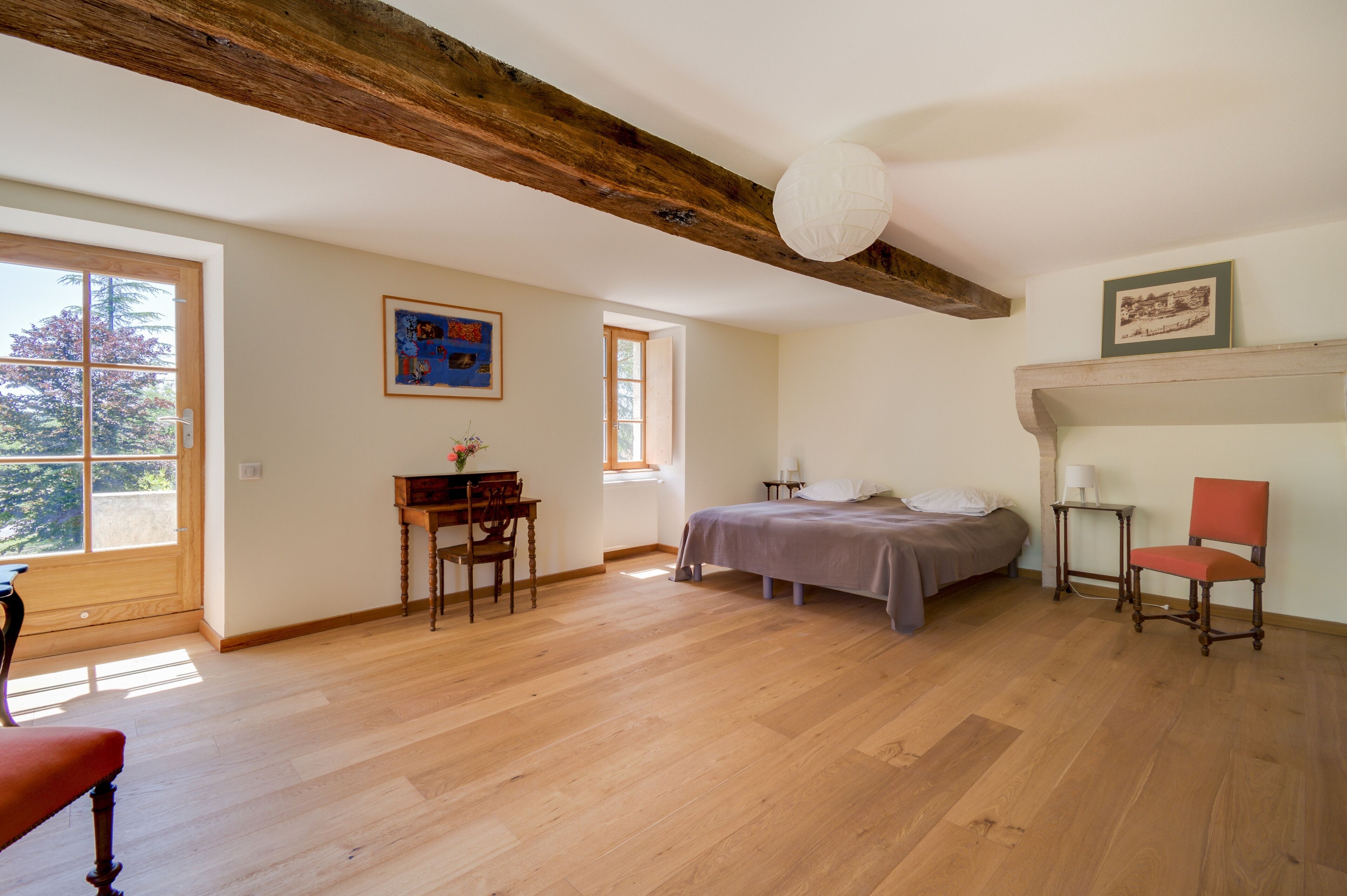 A spacious bedroom with a large bed, a wooden desk, a chair, and a small table. There is a window providing natural light and a view of trees outside.