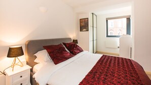 A neatly made bed with a white and red bedspread, a grey headboard, and two red pillows. There is a bedside lamp with a black shade on a white nightstand. A window with a view of a building is visible in the background. - Spacious 1 Bedroom Apartment in Kensington - A2 (London)