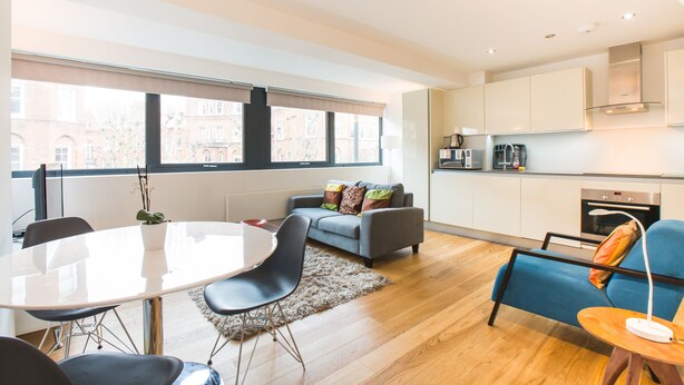 A modern apartment with a dining area, a sofa, a blue armchair, and a kitchen with white cabinets and a built-in oven. - Spacious 1 Bedroom Apartment in Kensington - A2 (London)