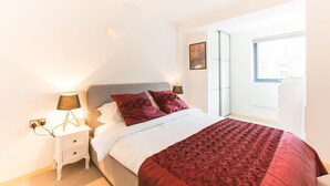 A bedroom with a bed, nightstand, lamp, and a window with a view. - Spacious 1 Bedroom Apartment in Kensington - A2 (London)