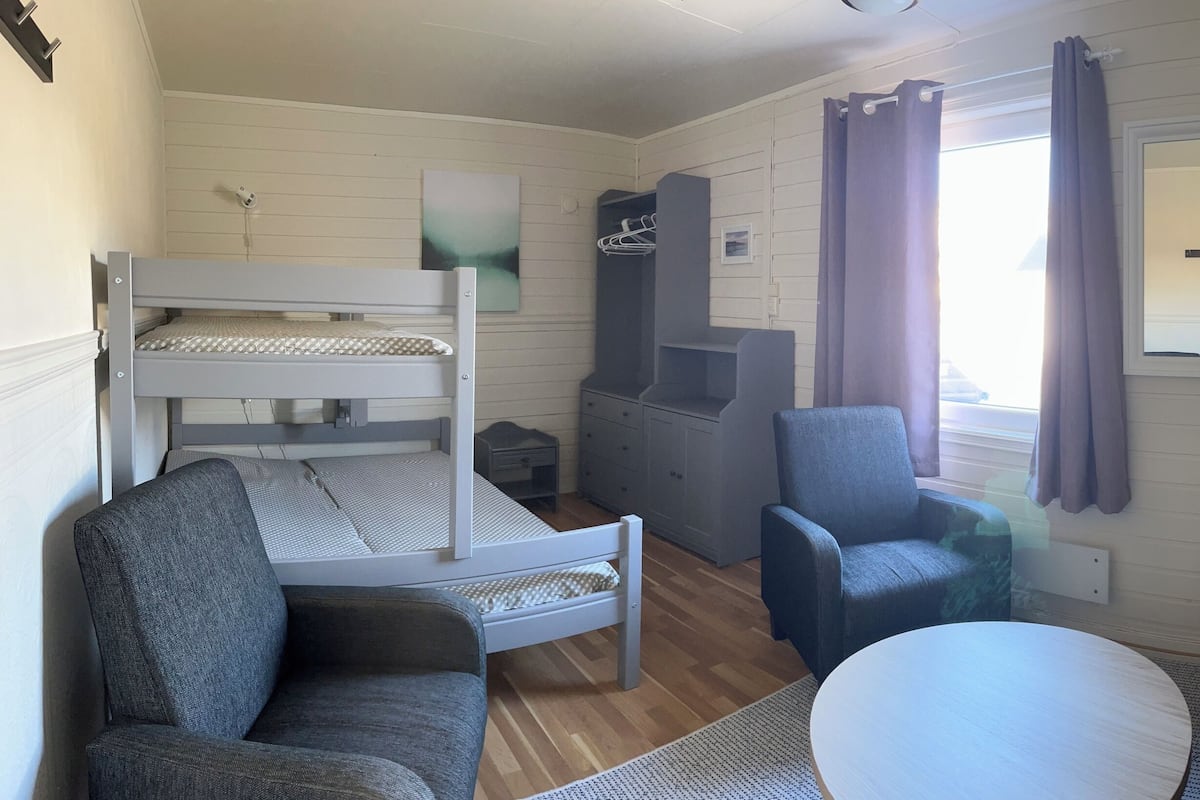 A room with a bunk bed, a chair, a sofa, a small table, and a window with curtains.