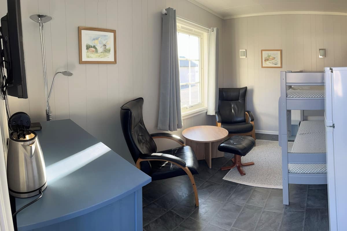 A compact room with a bunk bed, a desk with a kettle, a chair, a round table, and a window with curtains.