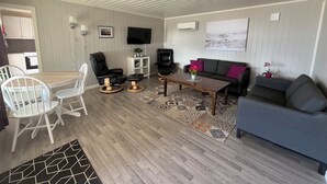 A living room with a sofa, chairs, a coffee table, a TV, and a kitchen area in the background. - Oppmyre Camping (Oksnes)