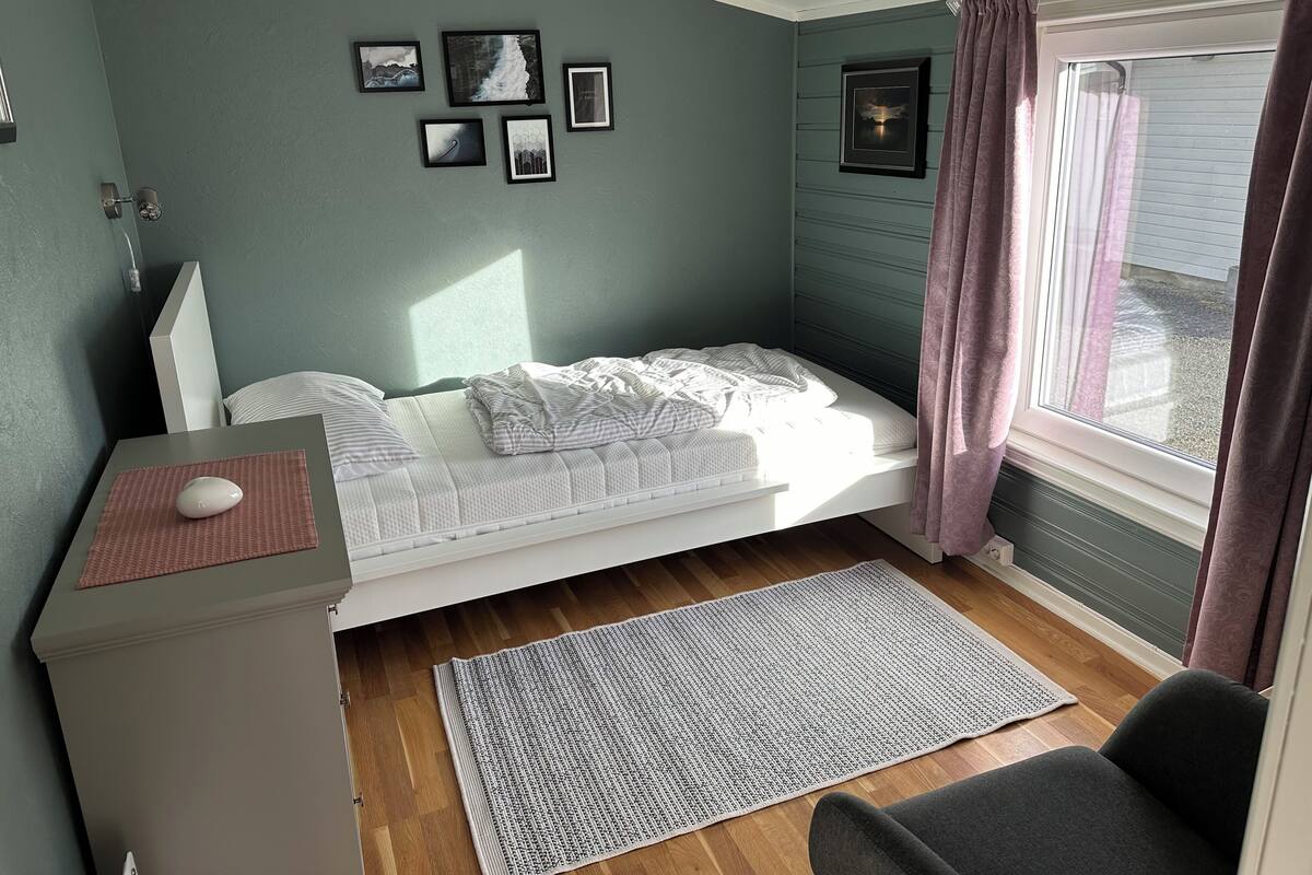 A small bedroom with a bed, a desk with a computer, a window with curtains, and several framed pictures on the wall.