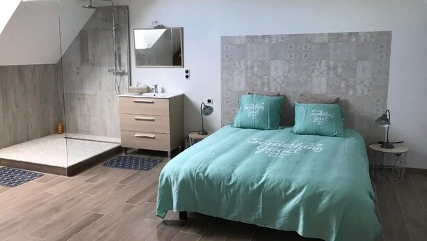 Een moderne slaapkamer met dakraam, een inloopdouche, een bed met blauwgroene beddengoed en een grote spiegel boven het wastafeltje.