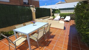 Une terrasse avec une table et des chaises, une petite table ornée d’un vase et un coin salon avec une nappe blanche.