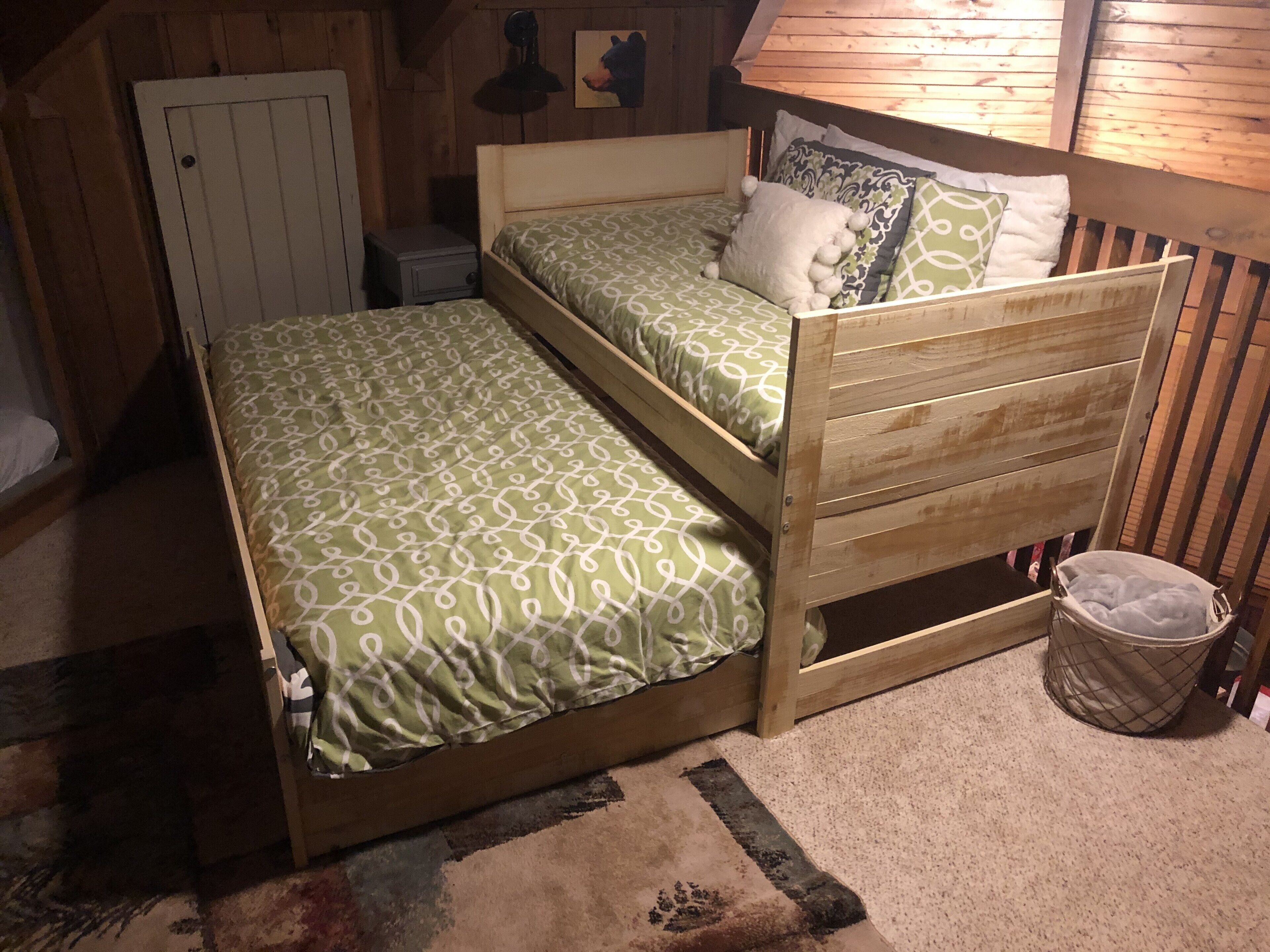 A room with two wooden bunk beds, a small bedside table, a lamp, and a trash bin.