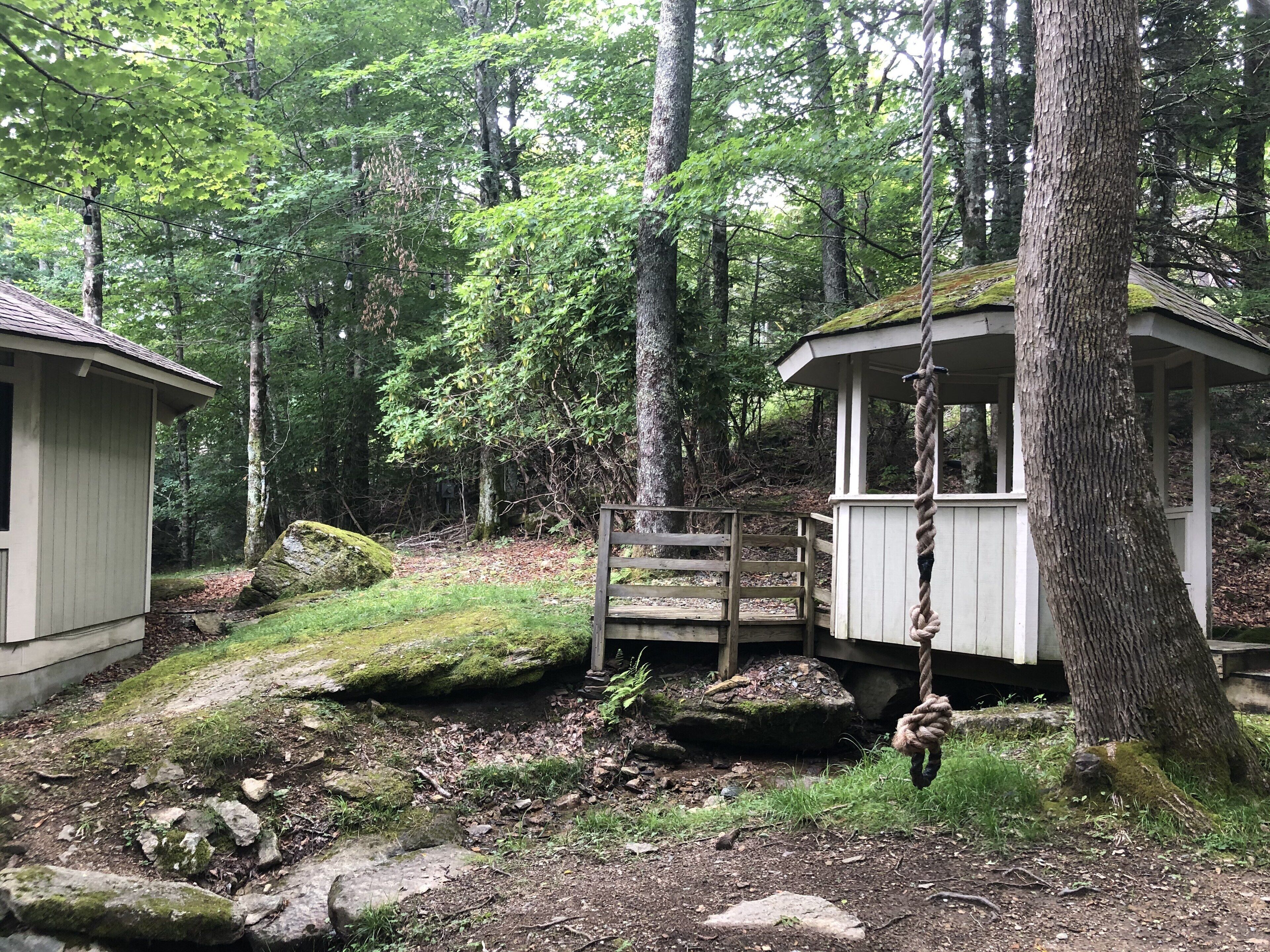 A rustic cabin with a wooden deck and a rope swing, surrounded by a forested area with moss-covered rocks and trees.