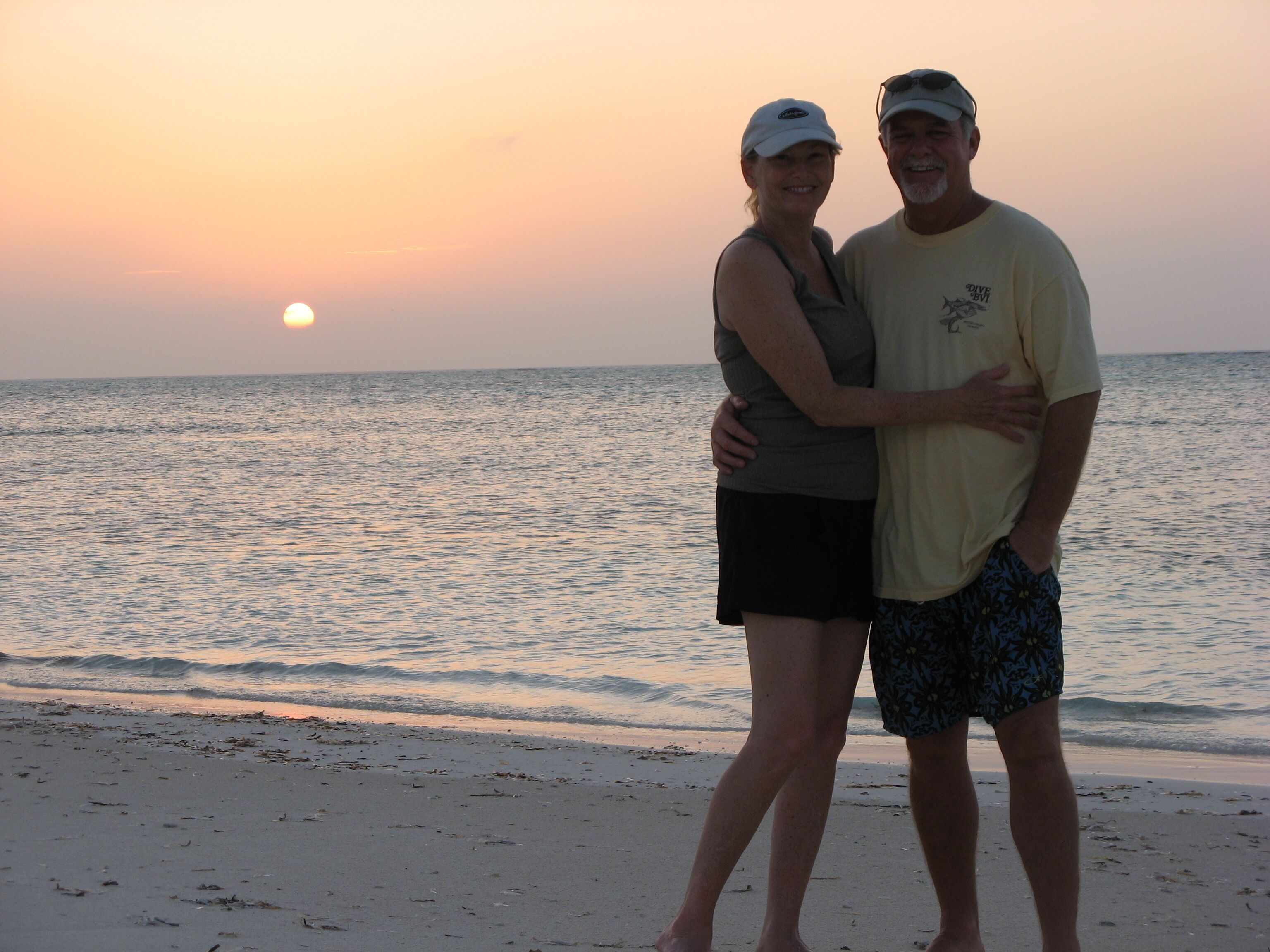 Ein Paar umarmt sich am Strand, im Hintergrund Meer und Sonnenuntergang.