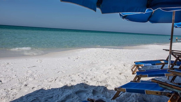 Una playa de arena blanca y un mar azul cristalino. Se ven sombrillas azules y sillas de madera para la playa.