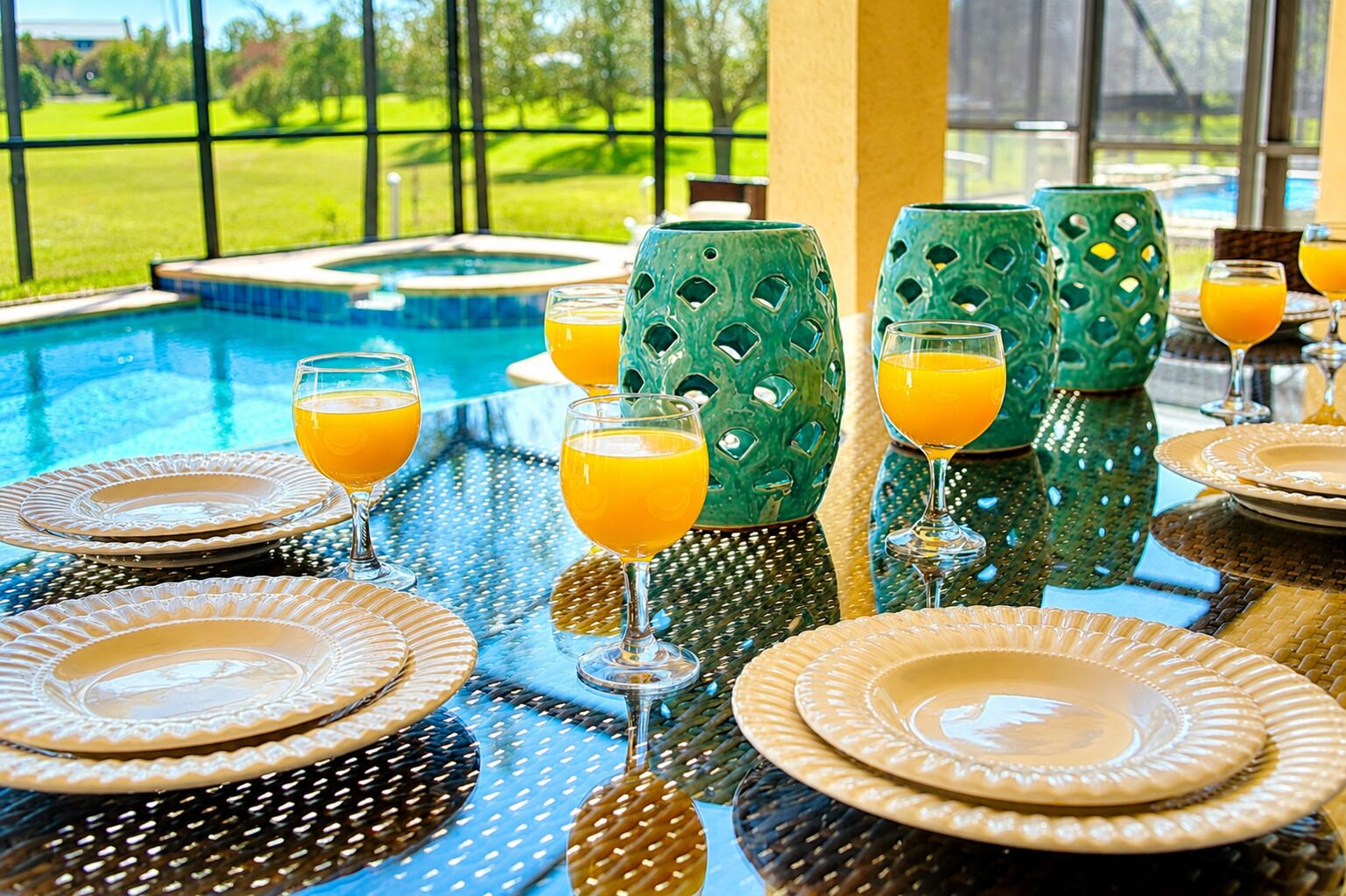 Mesa de comedor servida con platos, vasos y jugo de naranja, con vista a una piscina y un paisaje verde.