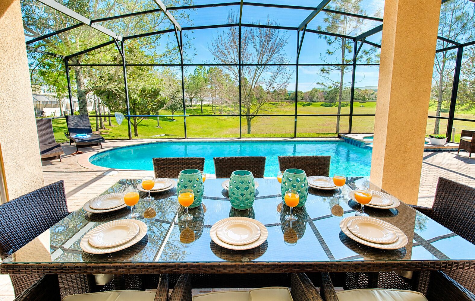 Un espacio de comedor al aire libre con una mesa de vidrio, sillas de mimbre y vistas a una piscina y follaje.