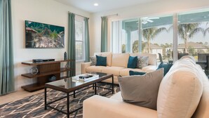 A modern living room with a beige sofa, a glass coffee table, and a TV mounted on the wall. - Delightful Home with Entertainment Loft at Encore (Kissimmee)