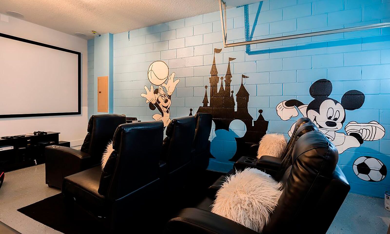 Een bioscoopzaal in huis met een groot scherm, zwarte leren loungestoelen en een muurschildering met tekenfilmfiguren.