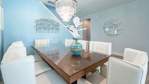 Zona pranzo con tavolo in legno, sedie imbottite bianche, un vaso blu con fiori bianchi, un lampadario e una decorazione geometrica a parete.
