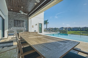 Un espacio moderno para comer al aire libre con piscina, pared de piedra y vista a un barrio residencial.