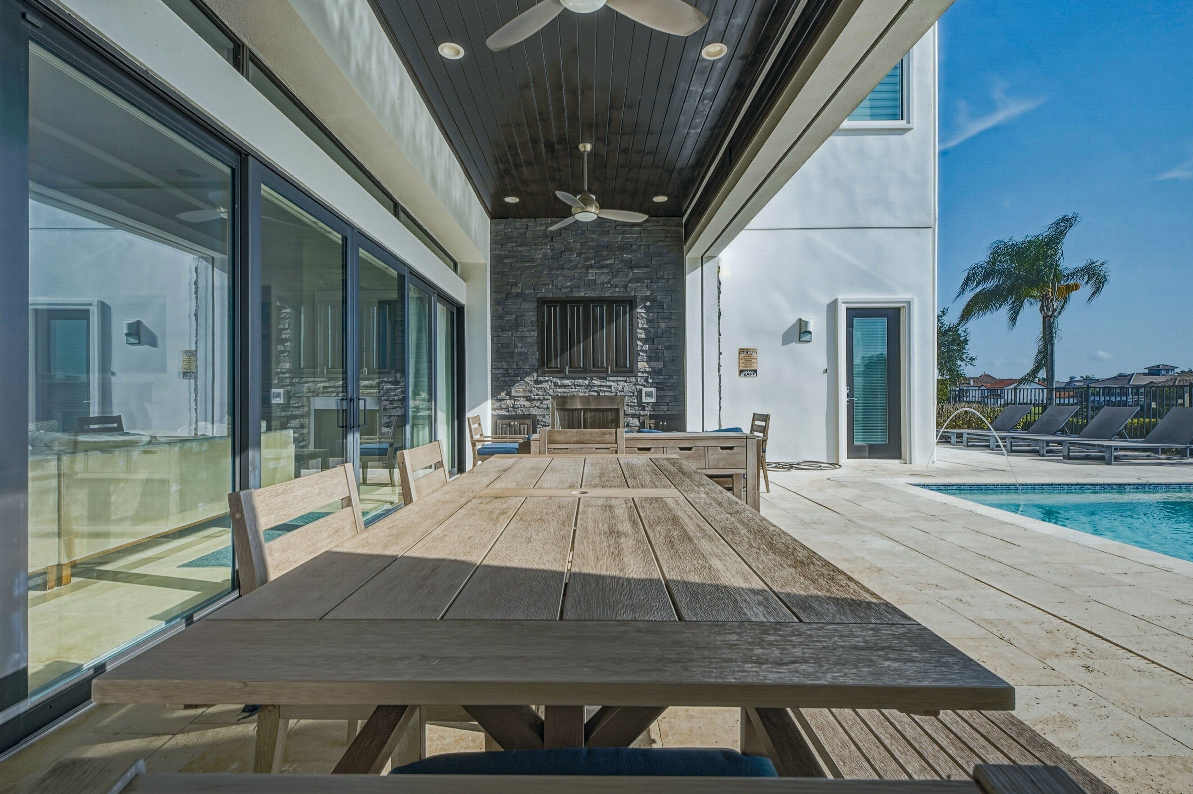 A wooden deck with a table and chairs, a stone wall, and a poolside area with lounge chairs.