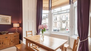 Um quarto com uma mesa de jantar de madeira, cadeiras, um banco, um vaso com flores, um abajur, um armário e uma janela com cortinas.