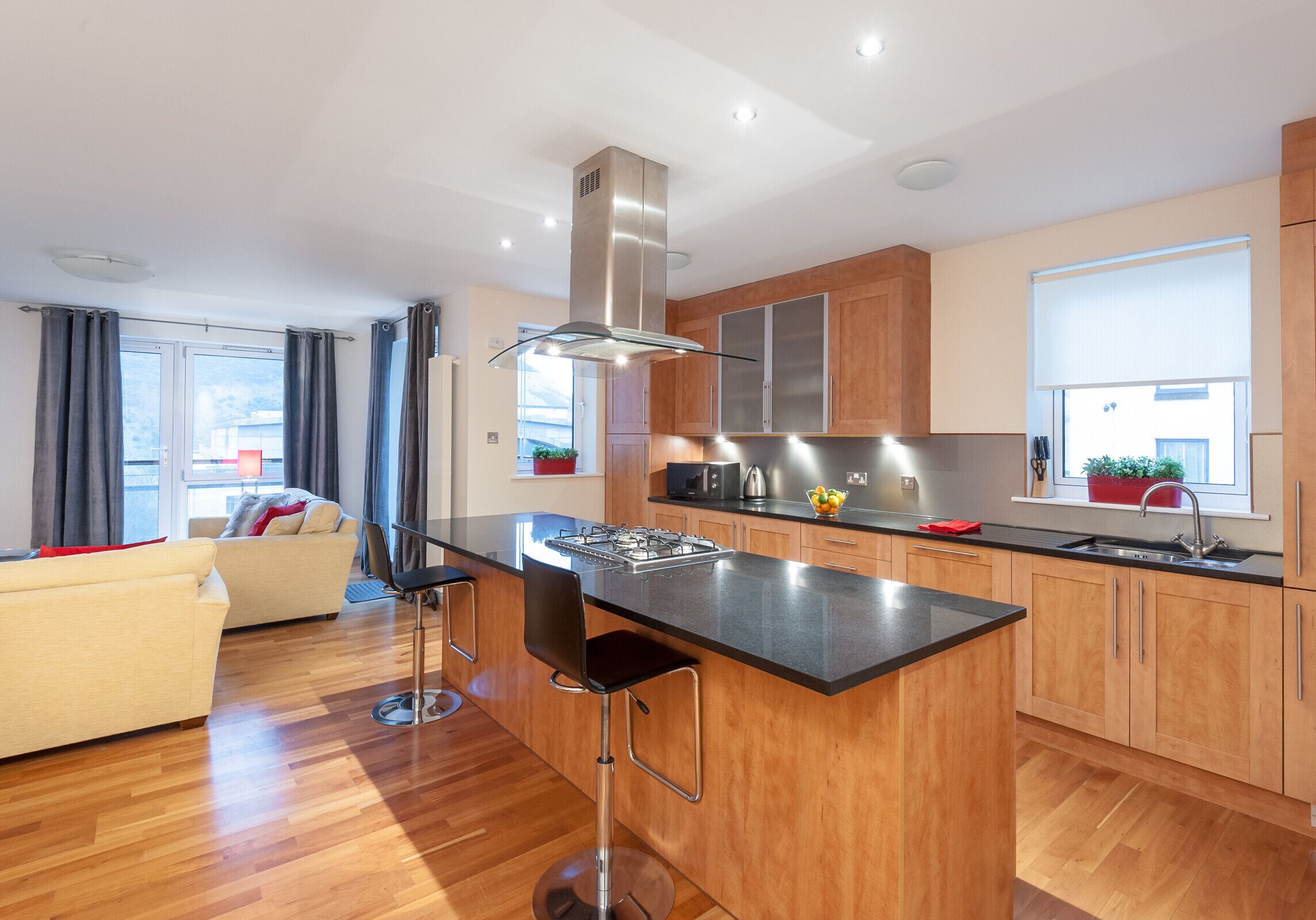 A modern kitchen with wooden cabinets, a central island with bar stools, a stainless steel range hood, and a window with a view.