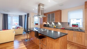 A modern kitchen with wooden cabinets, a central island with bar stools, a stainless steel range hood, and a window with a view. - Contemporary three-bedroom apartment with free parking space. Sleeps 5. (Edinburgh)