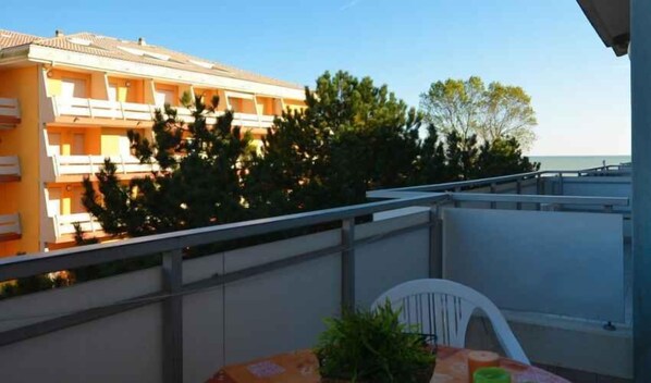 Un balcon donnant sur un immeuble et des arbres, une chaise blanche et une table recouverte d’une nappe.