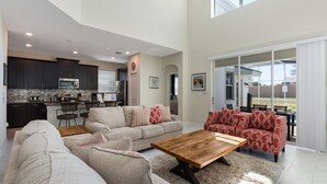 A spacious living room with a large sofa, a coffee table, and a red patterned sofa. A kitchen with dark cabinets and a microwave is visible in the background.