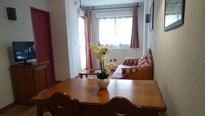Un coin repas avec une table et des chaises en bois, un téléviseur posé sur un meuble, une fenêtre avec des rideaux et un canapé à l’arrière-plan.