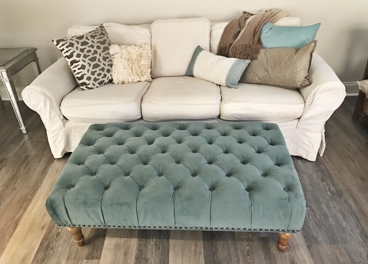 A tufted ottoman in teal fabric with a decorative border, placed in front of a white leather sofa with various cushions.