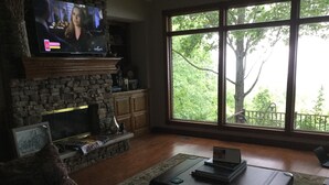 Un soggiorno con camino in pietra, televisione montata sopra e un'ampia finestra che si affaccia su alberi e un balcone.