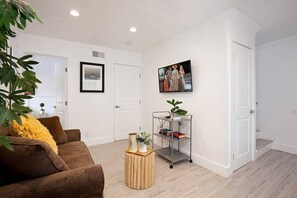 Un salon avec un canapé marron, une petite table d’appoint en bois, une étagère en métal avec des livres et une plante, et un tableau encadré au mur.
