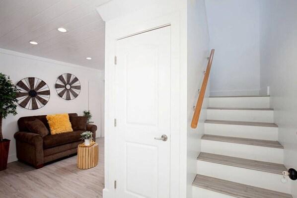 A modern living room with a brown sofa, a small wooden side table, and two wall decorations. A white door is open, leading to a staircase with wooden steps.